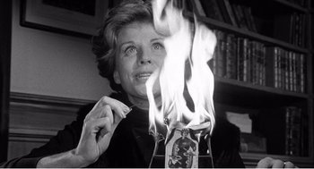 Movie still from “Night of the Eagle” (1962), directed by Sidney Hayers – An older woman is holding a candle in front of fire; Medium shot, Low angle