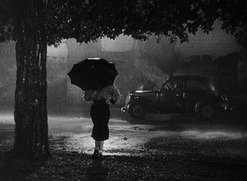 Movie still from “Nights of Cabiria” (1957), directed by Federico Fellini – A woman holding an umbrella under a tree in the rain; Wide shot, Over the shoulder angle