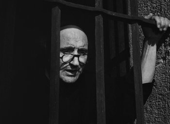 Movie still from “Nights of Cabiria” (1957), directed by Federico Fellini – An old man wearing glasses looking through the bars of a jail cell; Medium shot, Low angle