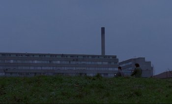 Movie still from “Nil by Mouth” (1997), directed by Gary Oldman – Two people are standing in a field near a building; Extreme Wide shot, Low angle