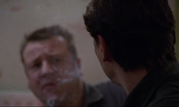 Movie still from “Nil by Mouth” (1997), directed by Gary Oldman – A man looking into a mirror while another man smokes a cigarette in front of him; Close Up shot, Over the shoulder angle