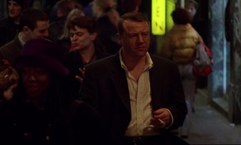 Movie still from “Nil by Mouth” (1997), directed by Gary Oldman – A man in a black suit and white shirt smoking a cigarette in a crowd; Medium shot, Over the shoulder angle