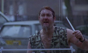 Movie still from “Nil by Mouth” (1997), directed by Gary Oldman – A man in a tank top is smoking a cigarette outside; Medium shot, Low angle