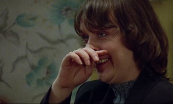 Movie still from “Nil by Mouth” (1997), directed by Gary Oldman – A young man with long brown hair is smiling and holding his finger to his mouth; Close Up shot, Over the shoulder angle