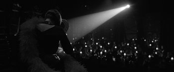 Movie still from “Nine” (2009), directed by Rob Marshall – A man in a black suit is standing in front of an audience; Medium shot, Over the shoulder angle