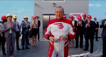 Movie still from “Nine Lives” (2016), directed by Barry Sonnenfeld – A man in a red and white racing suit holding a steering wheel; Medium shot, Over the shoulder angle