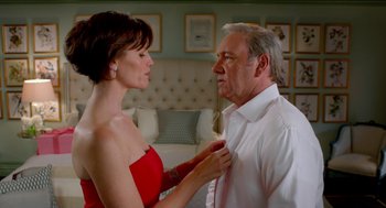 An older man adjusting a woman's neck tie in front of a bed