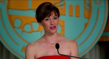 Movie still from “Nine Lives” (2016), directed by Barry Sonnenfeld – A woman in a strapless red dress speaking at a podium; Close Up shot, Over the shoulder angle