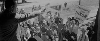 Movie still from “Nixon” (1995), directed by Oliver Stone – A crowd of people standing next to each other on a beach; Medium shot, High angle