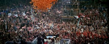 Movie still from “Nixon” (1995), directed by Oliver Stone – A crowd of people in an indoor arena with orange balloons overhead; Extreme Wide shot, High angle