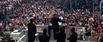 Movie still from “Nixon” (1995), directed by Oliver Stone – A crowd of people in a room with a man standing at a podium; Wide shot, High angle