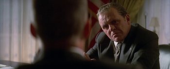 Movie still from “Nixon” (1995), directed by Oliver Stone – An older man wearing a suit and tie in front of an american flag; Close Up shot, Over the shoulder angle