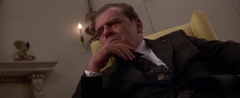 Movie still from “Nixon” (1995), directed by Oliver Stone – A man in a suit and tie sitting in a chair with his chin resting on his hand; Close Up shot, Low angle