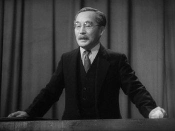Movie still from “No Regrets for Our Youth” (1946), directed by Akira Kurosawa – An old photo of a man in a suit and tie standing at a podium; Medium shot, Low angle