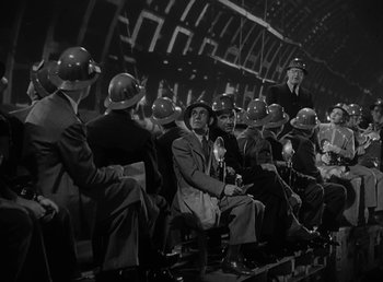 Movie still from “No Time for Love” (1943), directed by Mitchell Leisen – A black and white photo of a group of men sitting in a stadium; Wide shot, Low angle