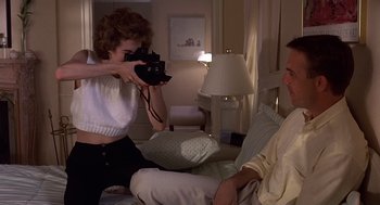 Movie still from “No Way Out” (1987), directed by Roger Donaldson – A woman taking a picture of a man sitting on a couch; Medium shot, Low angle