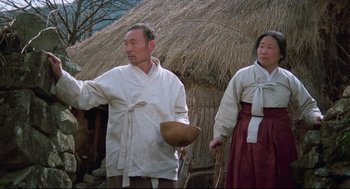 Movie still from “North Korean Partisan in South Korea” (1990), directed by Jeong Ji-yeong – A man and a woman standing next to each other in front of a thatched hut; Medium shot, Low angle