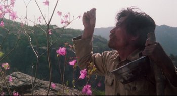 Movie still from “North Korean Partisan in South Korea” (1990), directed by Jeong Ji-yeong – A man standing next to a bush with pink flowers; Medium shot, Low angle