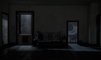 Movie still from “Nostalghia” (1983), directed by Andrei Tarkovsky – A person sitting on a bed in a dark room; Wide shot, High angle