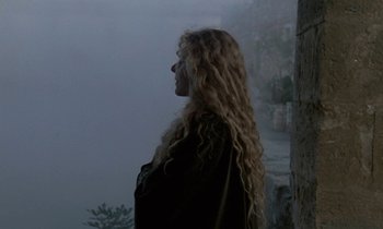 Movie still from “Nostalghia” (1983), directed by Andrei Tarkovsky – A woman with long blonde hair standing in front of a stone wall; Medium shot, Over the shoulder angle