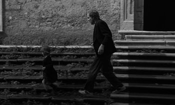 Movie still from “Nostalghia” (1983), directed by Andrei Tarkovsky – A man and a little girl are walking down steps; Wide shot, Low angle