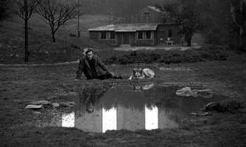 Movie still from “Nostalghia” (1983), directed by Andrei Tarkovsky – A man sitting on the ground next to a dog in front of a house; Wide shot, High angle