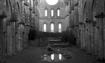 Movie still from “Nostalghia” (1983), directed by Andrei Tarkovsky – A person sitting on a bench in front of an old building; Extreme Wide shot, High angle