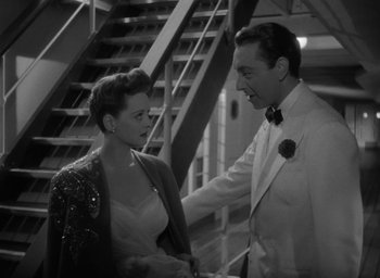 Movie still from “Now, Voyager” (1942), directed by Irving Rapper – A man and a woman in formal wear standing next to each other in front of a staircase; Medium shot, High angle