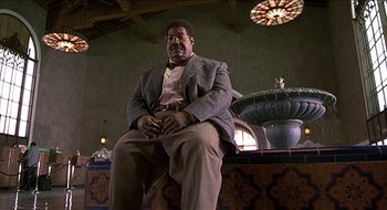 Movie still from “Nutty Professor II: The Klumps” (2000), directed by Peter Segal – A man in a suit sitting on a chair in front of a fountain; Medium shot, Low angle