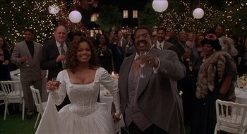 Movie still from “Nutty Professor II: The Klumps” (2000), directed by Peter Segal – A man and a woman are holding hands in front of a group of people; Medium shot, Low angle