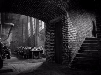 Movie still from “Oliver Twist” (1948), directed by David Lean – A group of people sitting at a table in front of a brick wall; Wide shot, Low angle