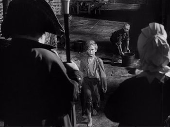 Movie still from “Oliver Twist” (1948), directed by David Lean – A young boy is standing in front of a group of people; Wide shot, High angle