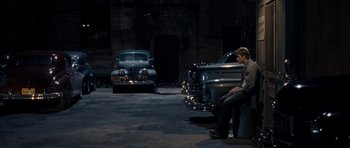 Movie still from “On the Road” (2012), directed by Walter Salles – A man sitting on top of a barrel next to two cars; Wide shot, Over the shoulder angle