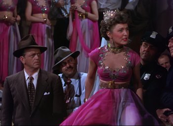 Movie still from “On the Town” (1949), directed by Gene Kelly – A woman in a pink dress is standing in front of a group of people; Medium shot, Low angle