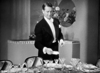 Movie still from “One Hour with You” (1932), directed by George Cukor – A black and white photo of a man standing at a dinner table; Medium shot, High angle