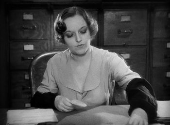 Movie still from “One Hour with You” (1932), directed by George Cukor – A woman sitting at a desk in front of a filing cabinet; Medium shot, High angle
