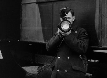Movie still from “One of Our Aircraft Is Missing” (1942), directed by Emeric Pressburger – A black and white photo of a man holding a camera; Medium shot, Low angle