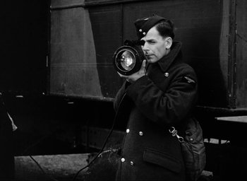 Movie still from “One of Our Aircraft Is Missing” (1942), directed by Emeric Pressburger – A black and white photo of a man holding a camera; Medium shot, Low angle
