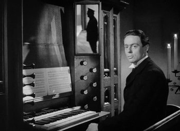 Movie still from “One of Our Aircraft Is Missing” (1942), directed by Emeric Pressburger – A black and white photo of a man in a suit and tie playing an organ; Medium shot, Low angle