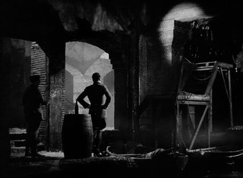 Movie still from “One of Our Aircraft Is Missing” (1942), directed by Emeric Pressburger – A man standing next to a barrel in an old building; Wide shot, Low angle