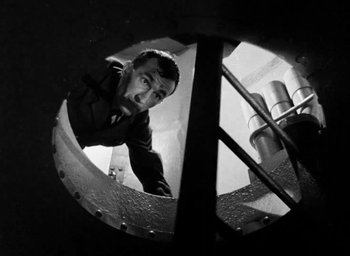 Movie still from “One of Our Aircraft Is Missing” (1942), directed by Emeric Pressburger – A black and white photo of a man looking through a hole in the wall; Medium shot, Overhead angle
