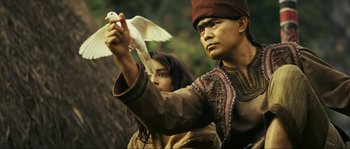 Movie still from “Ong Bak 2” (2008), directed by Tony Jaa – A man holding a white bird while standing next to a woman; Medium shot, Low angle