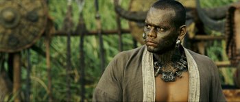 Movie still from “Ong Bak 2” (2008), directed by Tony Jaa – A man with a face tattoo is standing in front of an iron gate; Close Up shot, High angle