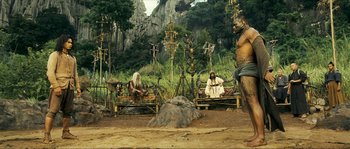 Movie still from “Ong Bak 2” (2008), directed by Tony Jaa – A group of people sitting on a bench in a field; Wide shot, Over the shoulder angle