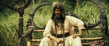 Movie still from “Ong Bak 2” (2008), directed by Tony Jaa – A man sitting in a chair wearing a turban; Medium shot, High angle