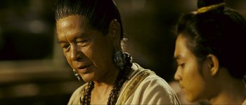 Movie still from “Ong Bak 2” (2008), directed by Tony Jaa – An older woman with long black hair wearing a white shirt; Close Up shot, Over the shoulder angle