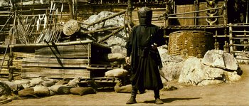 Movie still from “Ong Bak 2” (2008), directed by Tony Jaa – A man in a black outfit standing in front of a building; Wide shot, High angle
