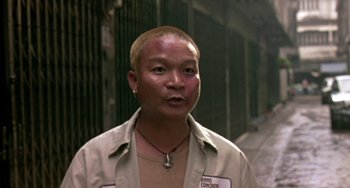 Movie still from “Ong-Bak: The Thai Warrior” (2003), directed by Prachya Pinkaew – A man in a tan shirt is standing in front of a building; Close Up shot, Low angle