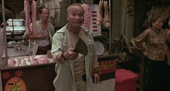 Movie still from “Ong-Bak: The Thai Warrior” (2003), directed by Prachya Pinkaew – A man holding a knife in his hand in front of other people in a market area; Medium shot, Over the shoulder angle