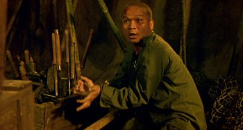 Movie still from “Ong-Bak: The Thai Warrior” (2003), directed by Prachya Pinkaew – A man sitting on a bench in front of some bamboo sticks; Medium shot, High angle